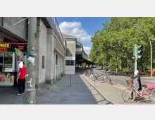 Eine Straßenszene in der Nähe einer U-Bahn-Station mit einem Döner-Imbiss auf der linken Seite. Im Hintergrund sind Schienen einer Hochbahn zu sehen, rechts stehen Fahrräder an einem Fahrradständer, umgeben von Bäumen und einer Straße.