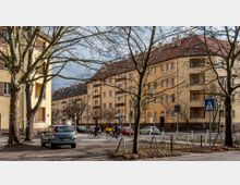 Straßenszene in einem Wohngebiet mit mehrstöckigen, gelb verputzten Wohnhäusern mit roten Ziegeldächern. Im Vordergrund stehen kahle Bäume, parkende Autos und ein Zebrastreifen, auf dem Personen mit Fahrrädern fahren.