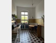 Helle Küche mit einem großen Fenster in der Mitte, das Tageslicht hereinlässt. Der Raum hat eine weiß-rot gemusterte hexagonale Fliesenboden, eine Holzarbeitsplatte, einen Gasherd auf der linken Seite und einen runden Holztisch mit Stühlen rechts. Die Fensterbank ist mit Topfpflanzen dekoriert.