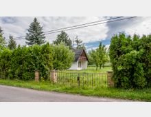 Kleines weißes Haus mit schwarzem Schrägdach, umgeben von dichter Vegetation, einschließlich hohen Bäumen und gepflegten Sträuchern. Im Vordergrund befindet sich ein schmiedeeiserner Zaun mit gemauerten Pfeilern entlang einer asphaltierten Straße.