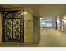 Unterführung mit karger Beleuchtung, grauen Wänden und Plakaten an den Seiten; links ein kunstvoll verziertes schmiedeeisernes Tor, rechts führen Treppen nach oben.
