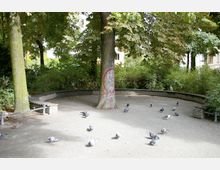 Ein kleiner, runder Platz inmitten eines Parks mit Kiesboden, einer geschwungenen niedrigen Steinmauer als Sitzgelegenheit und umgeben von Bäumen und dichtem Gebüsch. Eine große Kastanie steht zentral, mit etwas Graffiti bemalt, während mehrere Tauben auf dem Boden verteilt sind.