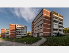 Mehrgeschossige Wohngebäude aus Beton mit roten Klinkerfassaden und Balkonen, umgeben von Rasenflächen, vereinzelten Bäumen und einem gepflasterten Gehweg. Der Himmel ist klar mit wenigen Wolken, und im Hintergrund sind weitere Bäume sichtbar.