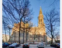 Backsteinkirche mit einem hohen Turm in neugotischem Stil, umgeben von unbelaubten Bäumen, die den Blick leicht verdecken. Im Vordergrund parken Autos auf einer Straße unter klarem, blauem Himmel.