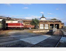 Ein historisches Bahnbetriebswerk mit einer roten Diesellokomotive und einem grünen Waggon auf den Gleisen. Im Hintergrund sind eine runde Drehscheibe, ein alter Ziegelfassade-Lokschuppen mit mehreren Toren sowie ein weißer Wasserturm mit spitzem Dach zu sehen.