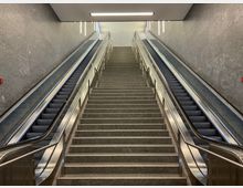 Eine breite Treppe aus Stein in einem modernen, schlicht gestalteten Innenraum wird von zwei Rolltreppen flankiert, die an den grauen Wänden mit metallischen Handläufen verlaufen. Oben an der Treppe befindet sich eine helle Beleuchtung und eine Decke mit ausgewogenen Linien.