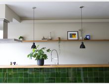 Moderne Küche mit einer Arbeitsplatte aus Holz und einer grünen Fliesenverkleidung. Über der Spüle mit einem hohen Wasserhahn hängen zwei schwarze Pendelleuchten, und im Hintergrund befinden sich eine schwebende Holzablage mit Dekorationsgegenständen und kleinen Pflanzen.