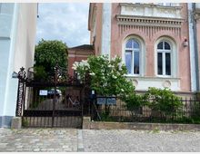 Rosa historische Hausfassade mit verzierten weißen Fensterumrandungen und zwei hohen Rundbogenfenstern, daneben ein schwarzes schmiedeeisernes Tor und Zaun mit dekorativen Mustern. Im Vordergrund befindet sich ein gepflasterter Gehweg, im Hintergrund ist ein kleiner begrünter Hof zu sehen.