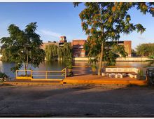 Holzplattform mit Sitzmöbeln aus Paletten und Pflanzkübeln, gelegen an einem Flussufer mit Blick auf alte Backsteingebäude und Trauerweiden am Wasser. Im Vordergrund gepflasterter Boden und ein Baum mit teils orangefarbenem Laub.