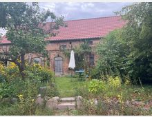 Backsteingebäude mit rotem Ziegeldach, umgeben von dichtem Garten mit Wildpflanzen, Bäumen und Blumen. Im Vordergrund steht ein grünes Rasenstück mit einem blauen Gartentisch, Stühlen und einem weißen Sonnenschirm.