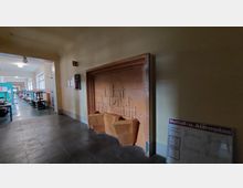 Ein Flur mit einem eingebauten Brunnen aus orangefarbenen Fliesen, verziert mit geometrischen Mustern. Im Hintergrund sind helle Fenster, Tische und Stühle eines Arbeitsbereichs zu sehen; an der Wand hängt ein Feuerlöscher sowie ein Notfall- und Alarmplan.