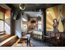 Ein gemütliches Café oder Bar mit hölzernem Interieur, einer Theke mit Barhockern und vereinzelten Tischen und Stühlen. An der rechten Wand befindet sich ein großes Wandbild mit stilisierten Figuren und gelben Strahlen, im Hintergrund teilweise freiliegendes Mauerwerk.
