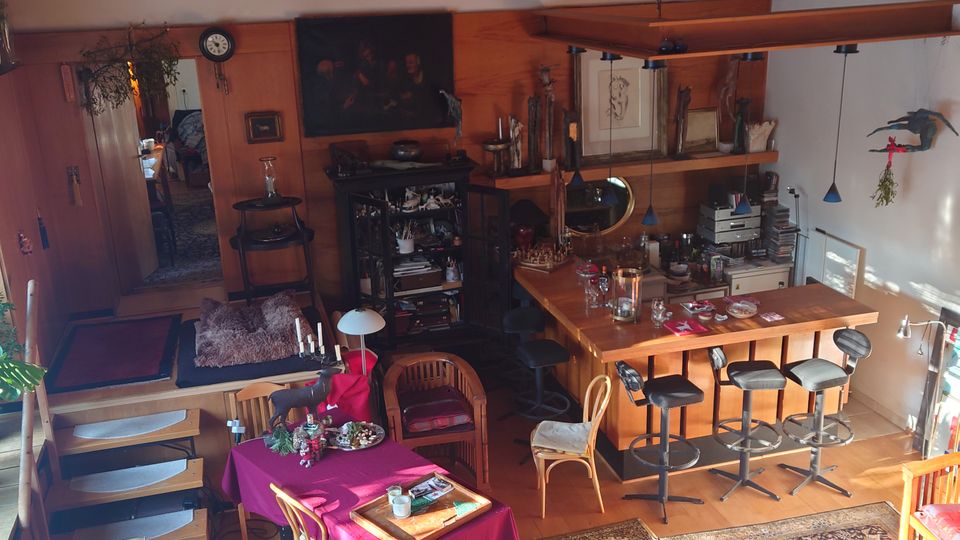 Wohnraum mit offenem Grundriss, Holzdach mit Rundbogen, gemusterten Teppichen und Holzböden. Der Raum umfasst eine Sitzecke, einen Esstisch mit roter Tischdecke, eine Küchenzeile mit Barhockern und Regale mit dekorativen Gegenständen.