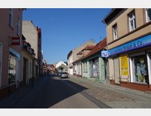 Schmale gepflasterte Straße in einer Kleinstadt mit zweistöckigen Wohn- und Geschäftshäusern. Auf der rechten Seite sind ein Reisebüro und ein verputztes Gebäude mit abblätternder Farbe zu sehen, links ein Gebäude mit LEGO-Schild und Schaufenstern.