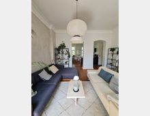 Modernes Wohnzimmer mit hellen Wänden und einem großen Teppich, auf dem ein schmaler Couchtisch steht. Links ein graues Sofa, rechts ein beiges Sofa, im Hintergrund weiße Backsteinwände und ein Durchgang zur Küche, dekoriert mit Pflanzen und Regalen. Eine große, runde Deckenleuchte hängt zentral im Raum.