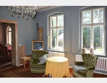 Wohnzimmer mit Holzboden, zwei grünen Polstersesseln und einem runden Tisch mit gelber Tischdecke vor großen Fenstern, die den Blick auf einen Garten mit Pavillon freigeben. Ein Kristallleuchter hängt von der Decke, und durch eine offene Tür ist ein Essbereich mit Holztisch und Stühlen sichtbar.