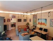 Innenraum eines offenen Atelier- oder Galerieraums mit Sofaecke und mehreren Tischen, an den Wänden hängen viele farbige Gemälde. Von der Decke verlaufen Kabel mit frei hängenden Lampen, im Hintergrund führt ein Durchgang in einen weiteren Raum.