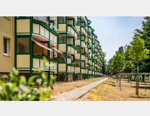 Mehrstöckiges Wohngebäude mit gelben Fassaden und grün gerahmten Balkonen, die mit Pflanzen und Markisen dekoriert sind. Im Vordergrund ein schmaler Gehweg, eine Rasenfläche mit Bäumen und Wäscheleinen, umgeben von einem parkähnlichen Bereich.