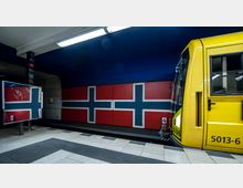 Untergrundbahnhof mit einer gelben U-Bahn auf der rechten Seite. Die Wand im Hintergrund ist mit einem rot-blau-weißen Kreuzmuster gestaltet, das an die norwegische Flagge erinnert.