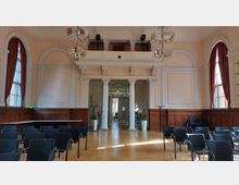 Ein historischer Saal mit hohen Decken, großen Fenstern mit roten Vorhängen und Holztäfelung an den Wänden. Im Vordergrund sind Stuhlreihen auf einem Parkettboden, im Hintergrund stehen weiße Säulen und eine Glastür, die zu einem weiteren Raum führt.