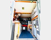 Innenraum eines umgebauten Fahrzeugs mit Wohnbereich. Sichtbar sind eine kleine Küche mit Hängeschränken, ein Etagenbett, mehrere Metallfächer und Stauraum, sowie eine auf den Boden gelegte blaue Matte.