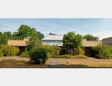 Flaches, modernes Gebäude aus hellen Ziegelsteinen mit einem zentralen Dach aus Metall, umgeben von Büschen, Bäumen und einer gepflasterten Fläche. Im Hintergrund sind hohe Bäume sichtbar, die eine parkähnliche Umgebung andeuten.