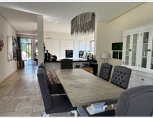 Esszimmer in einem modernen Wohnhaus mit einem rustikalen Esstisch aus Holz und gepolsterten grauen Stühlen. Im Hintergrund befindet sich eine offene, helle Küche mit weißen Einbauschränken, daneben ein weißer Vitrinenschrank und Blick durch eine Glastür auf den Garten.