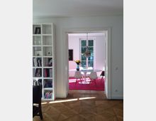 Innenansicht einer modernen Wohnung mit einem offenen Übergang zwischen zwei Räumen. Im Vordergrund ist ein Raum mit Parkettboden und einem weißen Bücherregal sichtbar; durch eine weiße Türöffnung führt der Blick in ein Esszimmer mit einem runden Tisch, weißen Stühlen, einem Kronleuchter und einem Fenster, das Tageslicht einlässt.