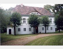 Zweistöckiges weißes Gebäude mit einem roten Ziegeldach und kleiner, bogenförmiger Gaube. Vor dem Gebäude befinden sich ein Rasenbereich, eine geschwungene Kiesauffahrt und mehrere Bäume, die den Eingangsbereich teilweise verdecken.