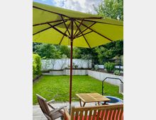 Privater Garten mit grünem Rasen, umgeben von einer weißen Mauer und Bepflanzung im Hintergrund. Im Vordergrund stehen Holzmöbel auf einer Steinterrasse unter einem großen, gelben Sonnenschirm.