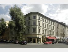 Straßenkreuzung mit einem mehrstöckigen Wohn- und Geschäftshaus im Gründerzeitstil an der Ecke. Die Fassade ist cremefarben mit symmetrischen Fenstern, im Erdgeschoss befinden sich Geschäfte, darunter ein Café und ein Ladengeschäft. Rechts ein U-Bahn-Zugang und parkende Autos entlang der Straße.