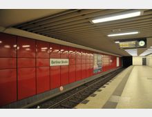U-Bahn-Station „Berliner Straße“ mit roten Wandpaneelen, Gleisen und Bahnsteig. Eine Uhr und eine Anzeigetafel mit Abfahrtsinformationen hängen unter der Decke, ergänzt durch Werbeplakate an den Wänden.