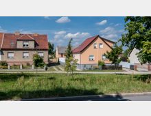 Straßenszene mit zwei Einfamilienhäusern, eines mit beiger Putzfassade und das andere mit grauem Verputz, beide mit roten Satteldächern. Davor liegt eine gepflegte Wiese mit jungen Bäumen, und ein Gehweg sowie eine asphaltierte Straße verlaufen im Vordergrund.