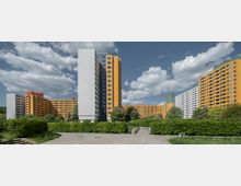 Blick auf eine Wohnsiedlung mit mehrstöckigen, orange- und weißfarbenen Plattenbauten, umgeben von Grünflächen, Gehwegen und Bäumen unter einem wolkigen Himmel. Im Vordergrund sind Büsche und ein gepflasterter Weg zu sehen.