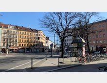 Straßenkreuzung in einem städtischen Viertel mit mehrstöckigen Altbauten in verschiedenen Farben, teils mit Geschäften im Erdgeschoss. Im Vordergrund stehen ein Kiosk mit Ziegeldach, mehrere Verkehrsschilder und geparkte Fahrräder, umgeben von Bürgersteigen und kahlen Bäumen.
