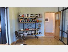 Ein Büro mit Holzboden und einer offenen Holztür, die zu einem Flur führt. An der Wand stehen Regale mit Ordnern, Büchern und Schreibtischutensilien; ein Stuhl und ein Ventilator befinden sich ebenfalls im Raum.