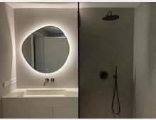 Modernes Badezimmer mit einem weißen Waschtisch und einem rundlich geformten, hinterleuchteten Spiegel an der linken Wand. Rechts befindet sich eine bodengleiche Dusche mit einer Regendusche und einer grauen Wandverkleidung.