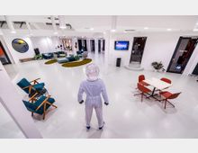 Ein moderner, offener Innenraum mit weißen Wänden und Böden, eingerichtet mit farbigen Sitzmöbeln, einem ovalen Teppich, Pflanzen und einem Tisch mit roten Stühlen. Im Vordergrund steht eine Person in einem futuristischen Raumanzug, während an den Wänden Fenster, Türen und ein Wandbildschirm sichtbar sind.