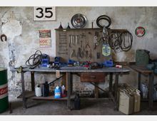 Eine Werkstatt mit einer alten Werkbank aus Holz und Metall, darauf Werkzeuge, Flüssigkeitsbehälter und verschiedene Kleinteile. An der Wand hängen geordnete Werkzeuge, Riemen und dekorative Schilder, vor einem Hintergrund aus rauem Putz.
