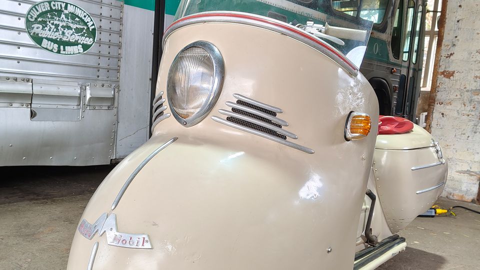 Vintage-Ausstellungsraum mit einem cremefarbenen, dreirädrigen Roller mit großer Windschutzscheibe im Vordergrund und einem grün-silbernen Linienbus im Hintergrund. Das Fahrzeug steht auf Betonboden unter einem Wellblechdach neben einer Backsteinwand mit Route-66-Schild.