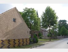 Straßenszene mit einer historischen Kirche aus Naturstein im Hintergrund, umgeben von Bäumen und einem kleinen gepflegten Garten. Im Vordergrund steht ein Gebäude mit Putzfassade und gemauertem Sockel.