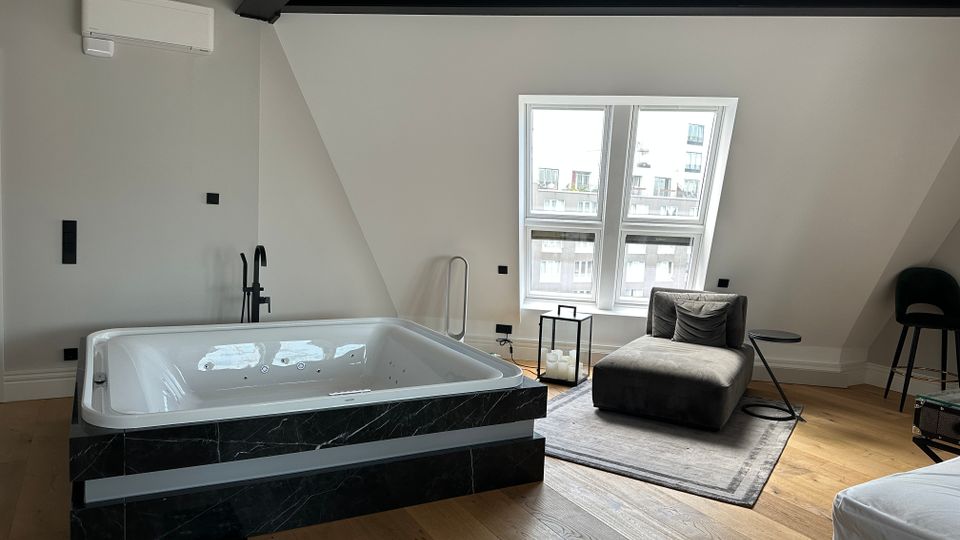 Modernes Dachgeschosszimmer mit freistehender Whirlpool-Badewanne in dunkler Marmorverkleidung, hellem Holzfußboden und sichtbaren schwarzen Deckenbalken. Neben einem Doppelfenster stehen eine graue Liege auf Teppich, ein kleiner Beistelltisch und eine Laterne mit Kerzen.