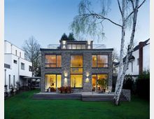 Moderne zweigeschossige Villa mit einer Fassade aus dunklem Backstein, großen Glasfenstern und einer Terrasse, umgeben von einem gepflegten grünen Rasen und Bäumen. Die Beleuchtung im Inneren des Hauses ist sichtbar und schafft einen warmen Kontrast zur Dämmerung.