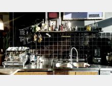 Ein Café-Thekenbereich mit glänzenden schwarzen Fliesen, einer Edelstahl-Arbeitsfläche und einer Espressomaschine auf der linken Seite. Darüber befindet sich ein Regal mit Gläsern, Flaschen und Küchenutensilien, während ein Doppelwaschbecken mit einem hohen Wasserhahn auf der rechten Seite sichtbar ist.