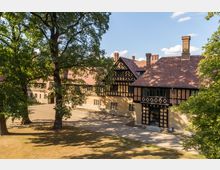 Historisches Herrenhaus im Tudorstil mit Fachwerkelementen und roten Ziegeldächern, umgeben von großen, schattenspendenden Bäumen auf einem gepflasterten Vorplatz bei sonnigem Wetter.