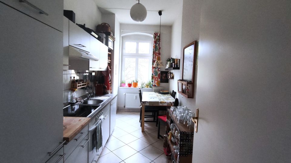 Narrow galley kitchen with white cabinets and a long countertop with sink and stove along the left wall, leading to a small dining table near a large window. The room has light tile flooring, pendant lights, potted plants on the windowsill, and a side shelf with glassware and a wall-mounted board on the right.