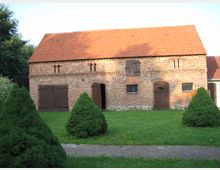Backsteinbau im ländlichen Stil mit Satteldach aus roten Ziegeln, mehreren Holztüren und kleinen Fenstern. Im Vordergrund ein gepflegter Rasen mit symmetrisch angeordneten Buchsbaumsträuchern.