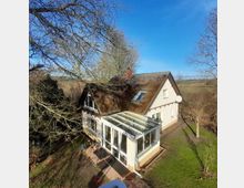 Haus mit Reetdach in ländlicher Umgebung, mit einem Wintergarten aus Glas und einer Außenterrasse. Umgeben von kahlen Bäumen und Wiesen an einem sonnigen Tag.