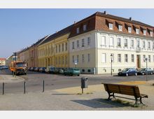 Straßenszene mit historischen Gebäuden in Pastelltönen, gepflasterter Fahrbahn und geparkten Autos. Im Vordergrund ein Bürgersteig mit einer Holzbank und einem Mülleimer, rechts ein Straßenschild und links ein orangefarbener Lkw.