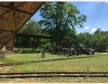 Industriegelände mit Gleisen im Vordergrund, einem alten Mauergebäude links, und einer Förderanlage aus Stahl und Maschinen auf einer Wiese, umgeben von Bäumen und Grünfläche. Ein rostiger Laufsteg aus Stahl erstreckt sich im Hintergrund.